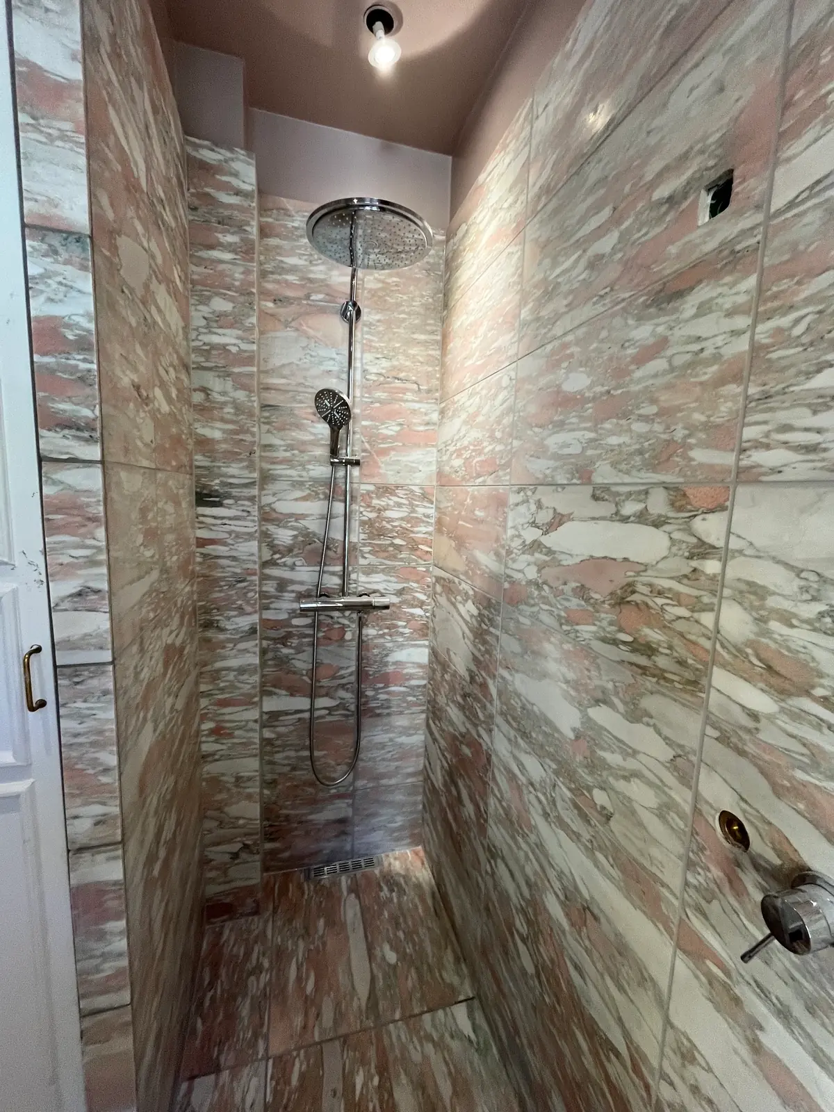 Bruseniche beklædt fra gulv til loft med rosa marmorfliser med kraftig åretegning, med regnbruser på væggen, en hylde i samme sten og glasvæg ind mod rummet