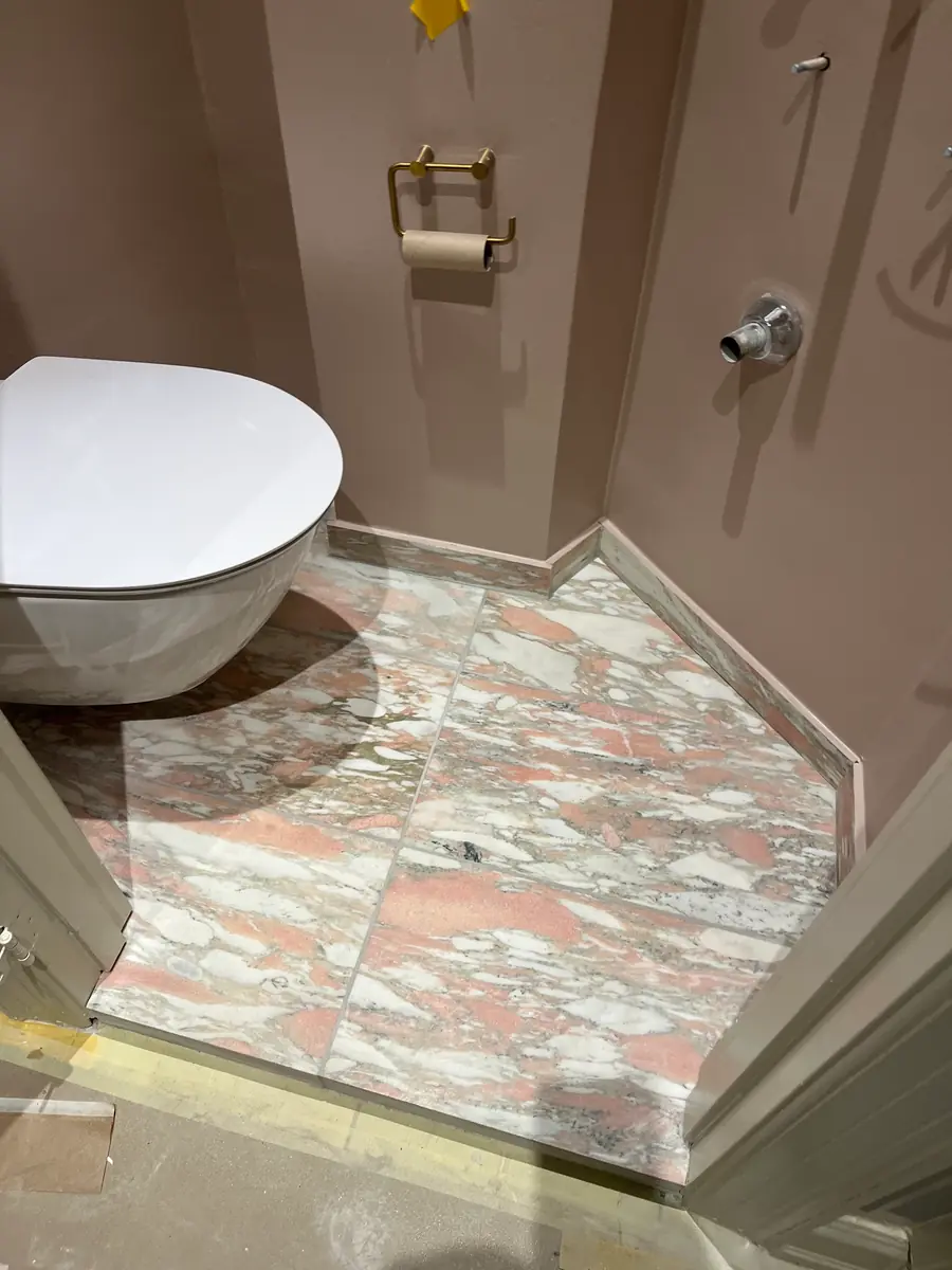 Toiletkrog med nyt gulv i rosa marmorfliser lagt helt ind i hjørnet og en sokkelkant i samme sten, mod støvet lilla vægge