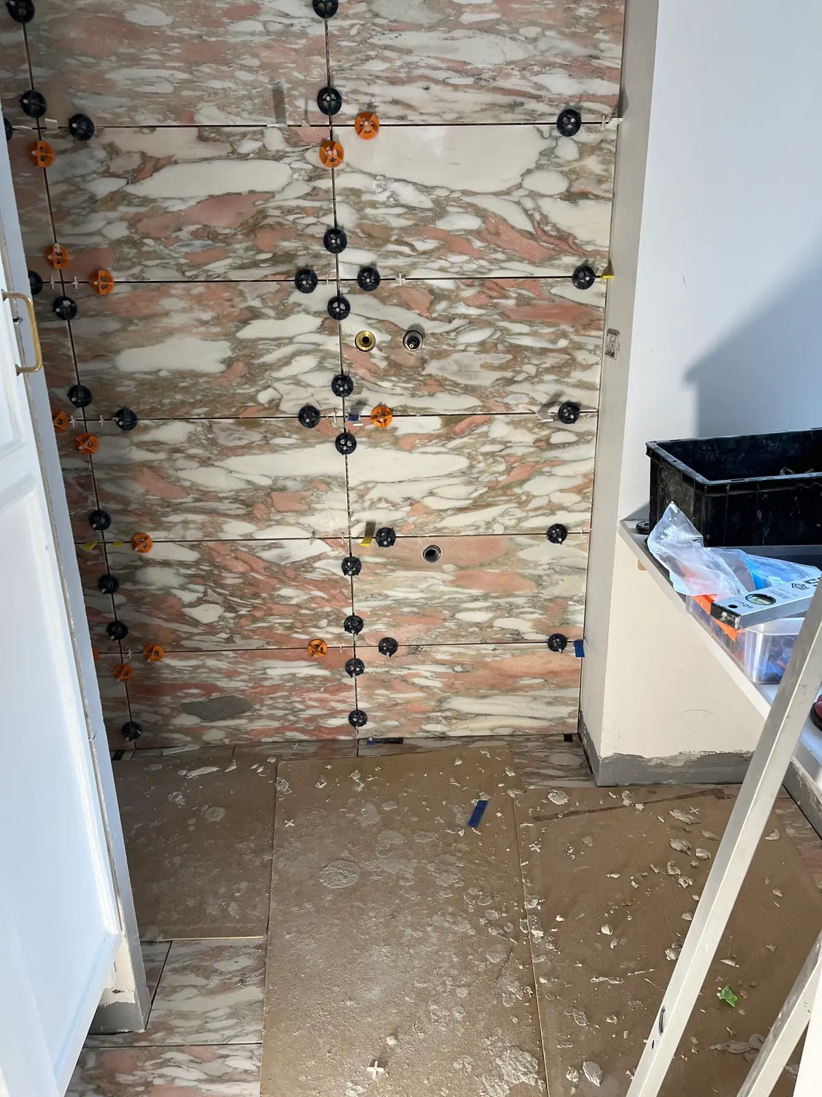 Rosa marmorvægfliser sat op i fuld højde med sorte og orange nivelleringsklips i hver fuge og udsparinger til armatur