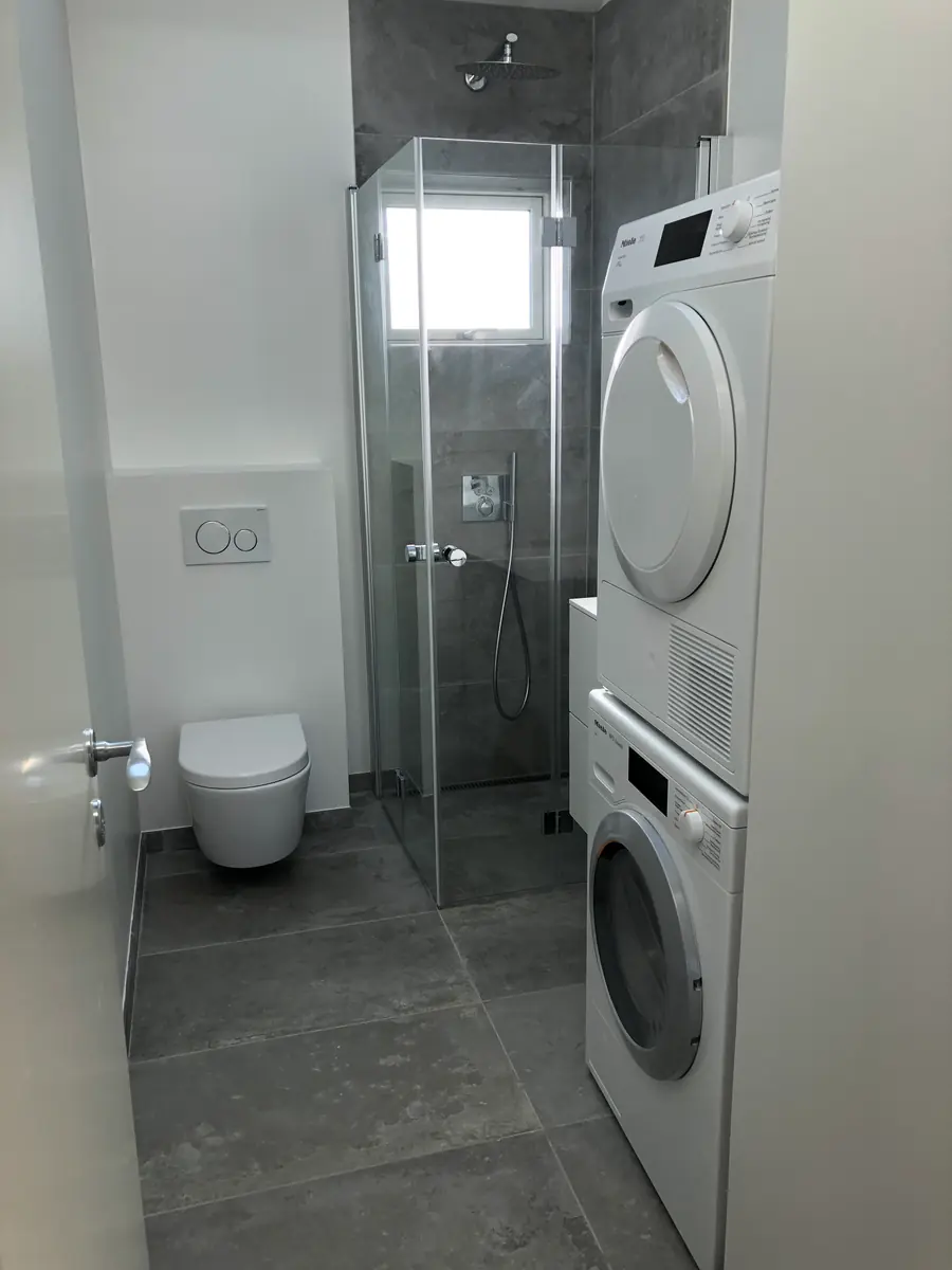 Smalt badeværelse med gråt flisegulv og grå storformatfliser i brusekabinen bag glasdøren, med væghængt toilet og en vaskesøjle med vaskemaskine og tørretumbler