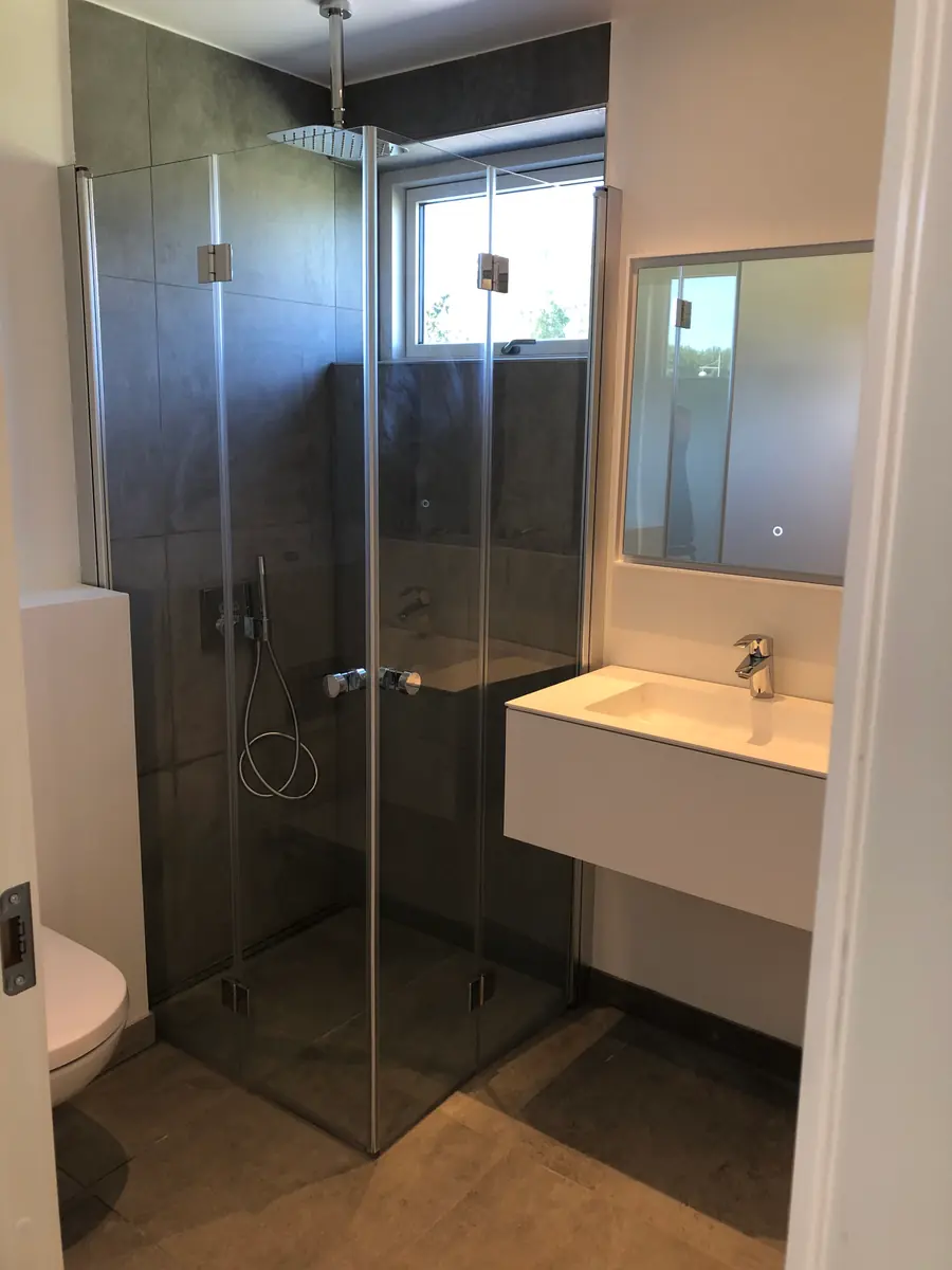 Færdigt badeværelse med mørkegrå storformatfliser, hjørnebruser i glas, hvid vask og væghængt toilet