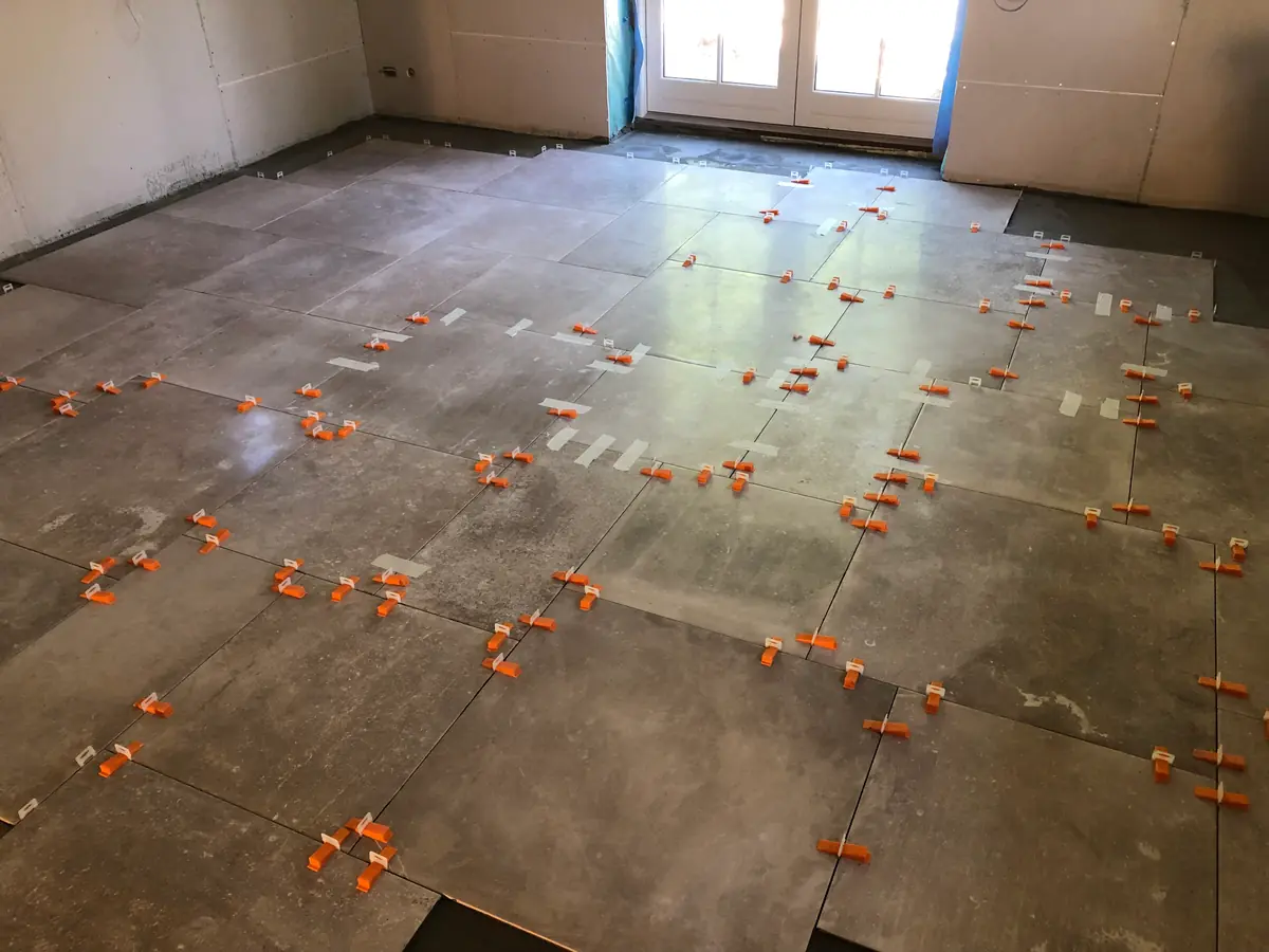 Store gråmelerede gulvfliser lagt i stort rum med orange nivelleringskiler i alle fuger og terrassedør bagest