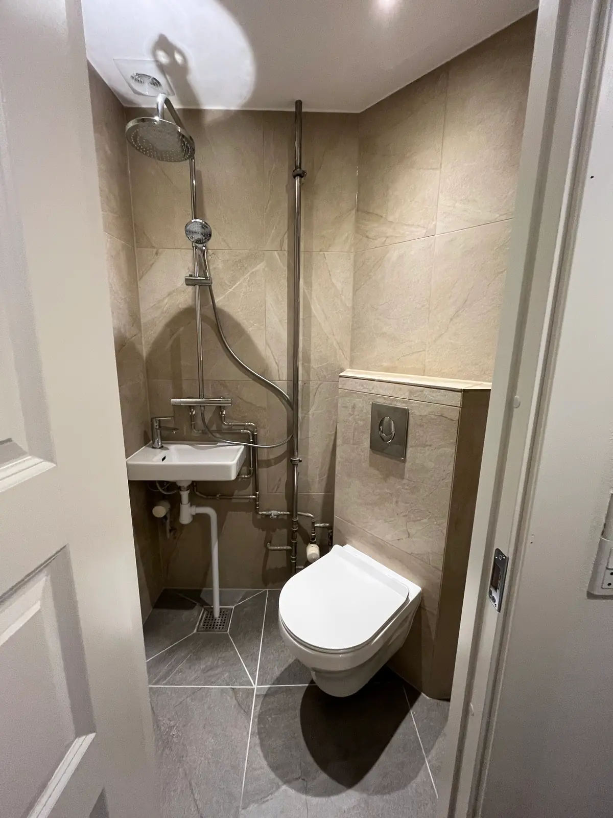 Færdigt minibad set fra døren med beige storformatfliser på væggene, gråt flisegulv lagt på diagonalen og indbygningsvæggen bag toilettet beklædt med fliser