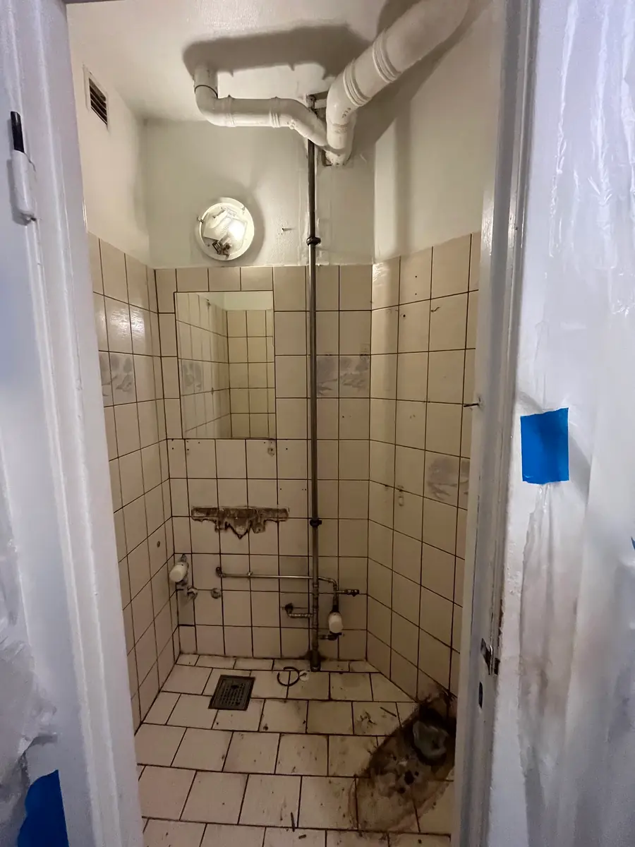 Nedslidt bruserum med cremefarvede firkantfliser, dekorfliser, spejl, gulvafløb og tæret rørføring set fra døren